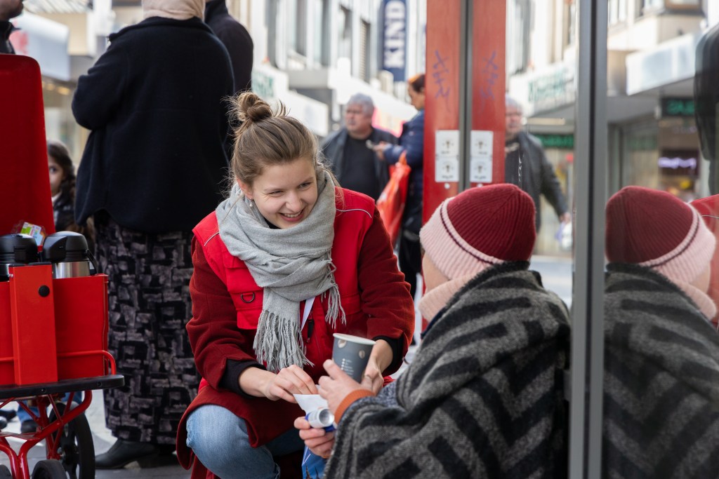 Eine ehrenamtliche Helferin des Kaffee-und-Knifte-Teams gibt einer auf dem Boden sitzenden Person ein Getränk.
